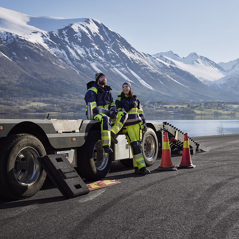 Two workers in high-visibility workwear taking a break by a transport vehicle, set against a mountainous landscape—designed for visibility, comfort and reliability on the job.