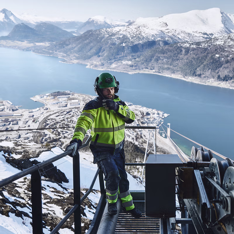 High-altitude industrial work in harsh winter conditions, featuring a worker in high-visibility protective clothing climbing a metal platform above a fjord, with snow-covered mountains in the background.