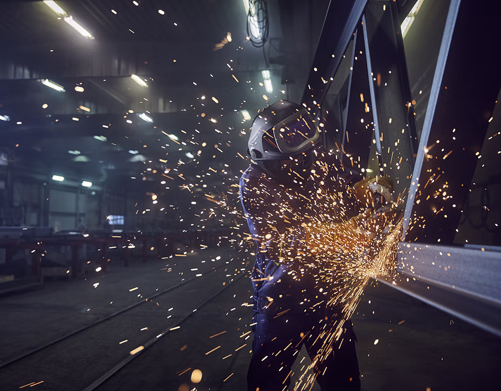 Industrial worker wearing flame-resistant protective clothing and a welding helmet, grinding a steel beam inside an industrial facility with sparks flying.