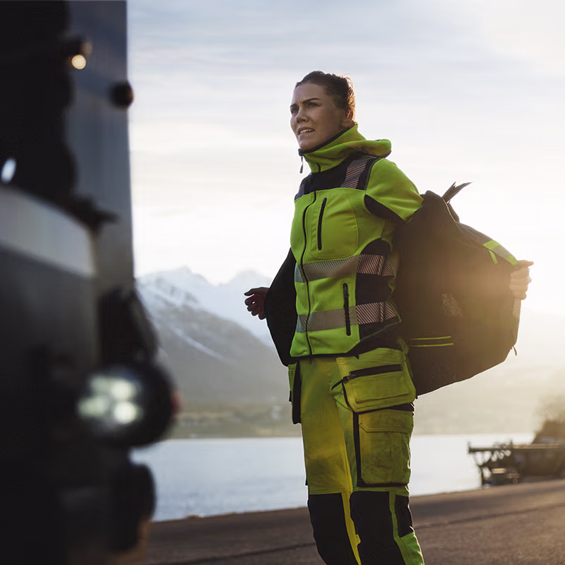 Worker in high-visibility workwear adjusting outer layer beside a transport vehicle at a roadside location—designed for visibility, flexibility and reliable performance in changing conditions.
