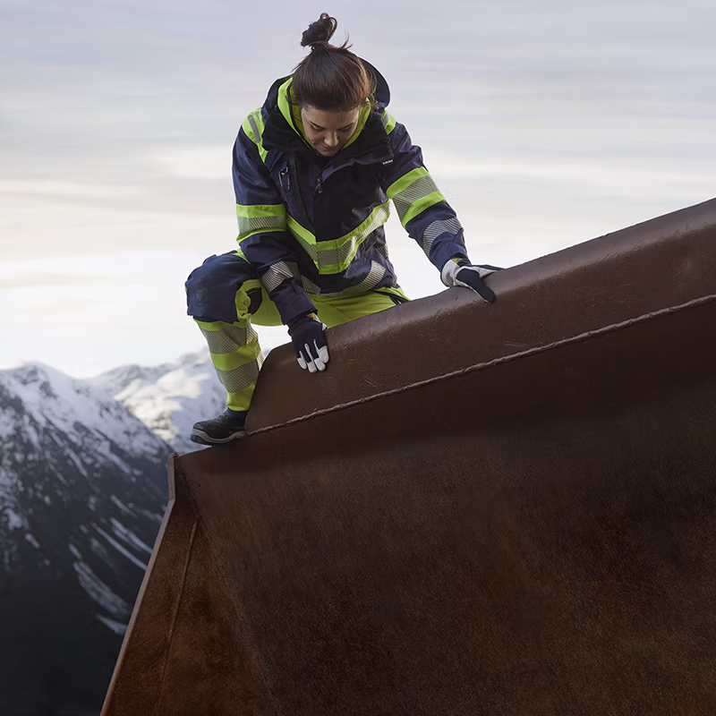 Worker in high-visibility workwear climbing onto heavy steel structure in a mountainous environment—built for mobility, durability and reliable protection in demanding conditions.