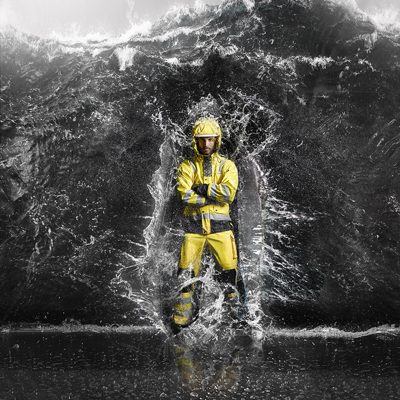 Worker in high-visibility rainwear standing firm as powerful waves crash around the body—engineered to withstand extreme weather, heavy rain and the toughest working conditions.