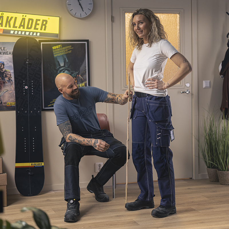 Two people indoors, one measuring the fit of Blåkläder work trousers worn by another person, with Blåkläder Workwear branding and wall posters visible in the background.