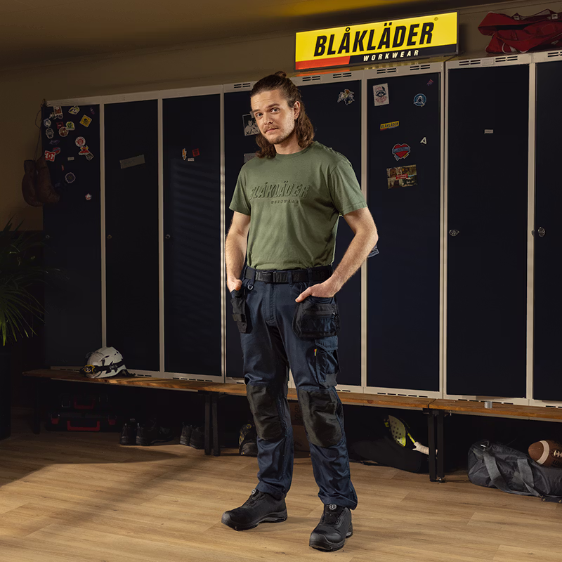 Person wearing Blåkläder workwear standing in a locker room, with dark lockers, a bench, and a Blåkläder Workwear sign visible in the background.