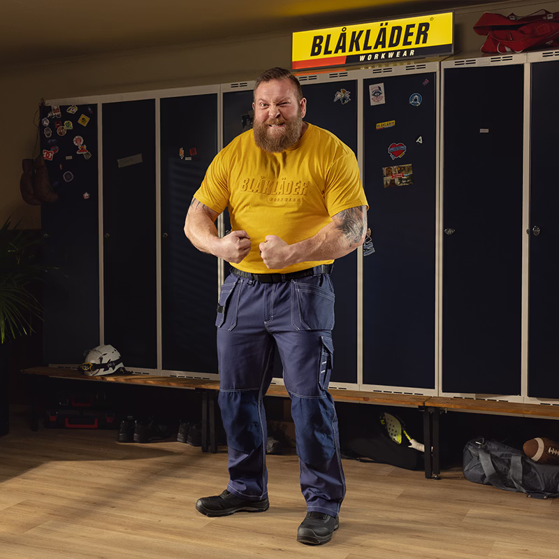 Person wearing a yellow Blåkläder work T‑shirt and blue work trousers standing in a locker room, with dark lockers, a bench, and a Blåkläder Workwear sign in the background.