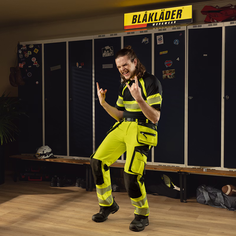 Person wearing high‑visibility Blåkläder workwear standing in a locker room, with dark lockers, a bench, and a Blåkläder Workwear sign in the background.