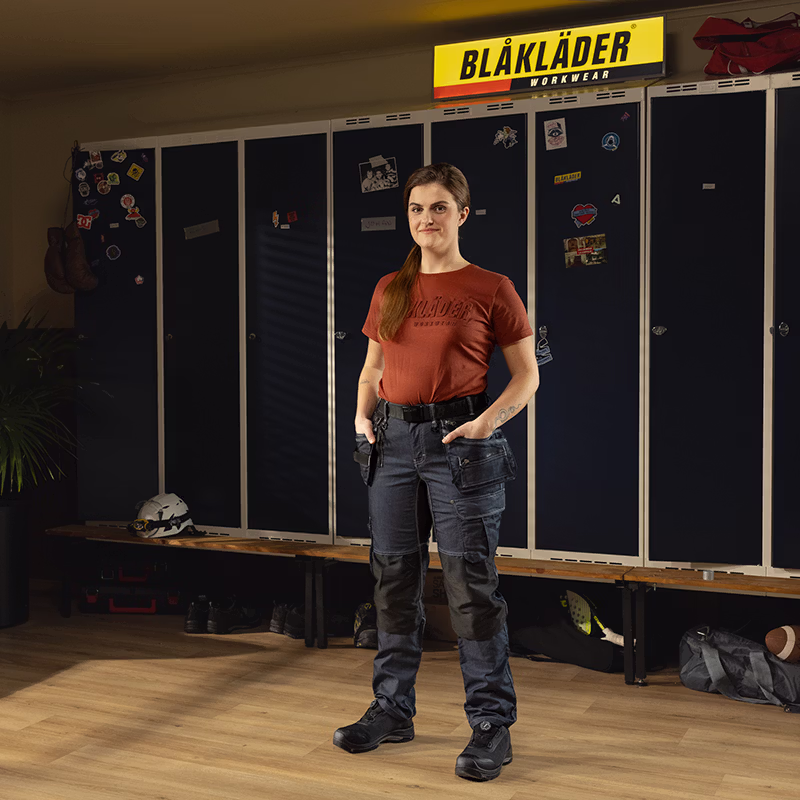 Person wearing a Blåkläder work T‑shirt and dark work trousers standing in a locker room, with dark lockers, a bench, and a Blåkläder Workwear sign in the background.