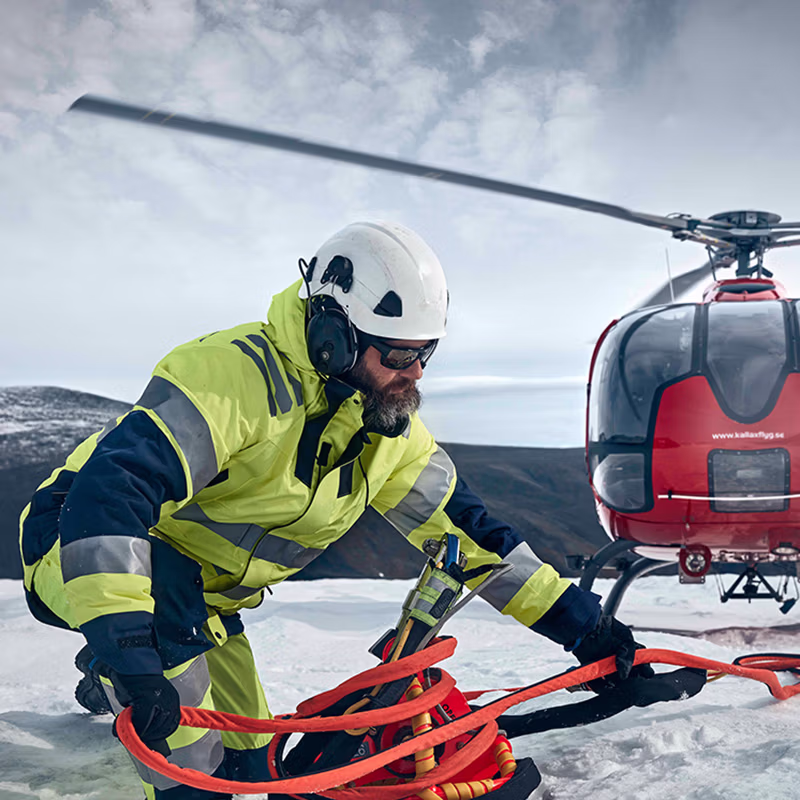 Worker in high-visibility winter workwear preparing equipment beside a helicopter in a snowy, exposed environment, built for demanding conditions and cold-weather safety.